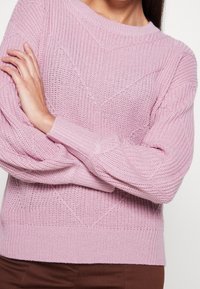 Rosa stickad tröja med texturerade mönster, rundad halsringning och ribbade muddar. Har avslappnade ärmar och en geometrisk design.