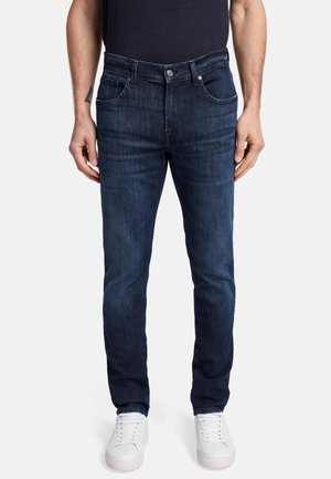 Dark wash denim jeans with a straight fit, featuring a button fly, five pockets, and subtle fading. Worn with white sneakers.