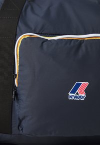 Tumši zils ceļojuma soma ar strukturētu audumu, kurai ir balts rāvējslēdzis, oranži akcenti un K-Way logo uz priekšējā kabatas.