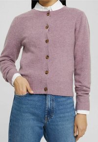 Kvinna som bär en ljuslila knappad kofta över en vit skjorta med krage och blå jeans, med handen i fickan.