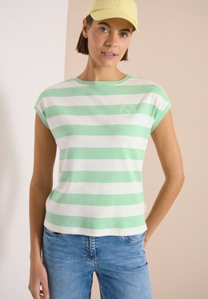 Jeune femme portant un débardeur rayé vert et blanc, un jean bleu et une casquette de baseball jaune clair, debout devant un fond beige.