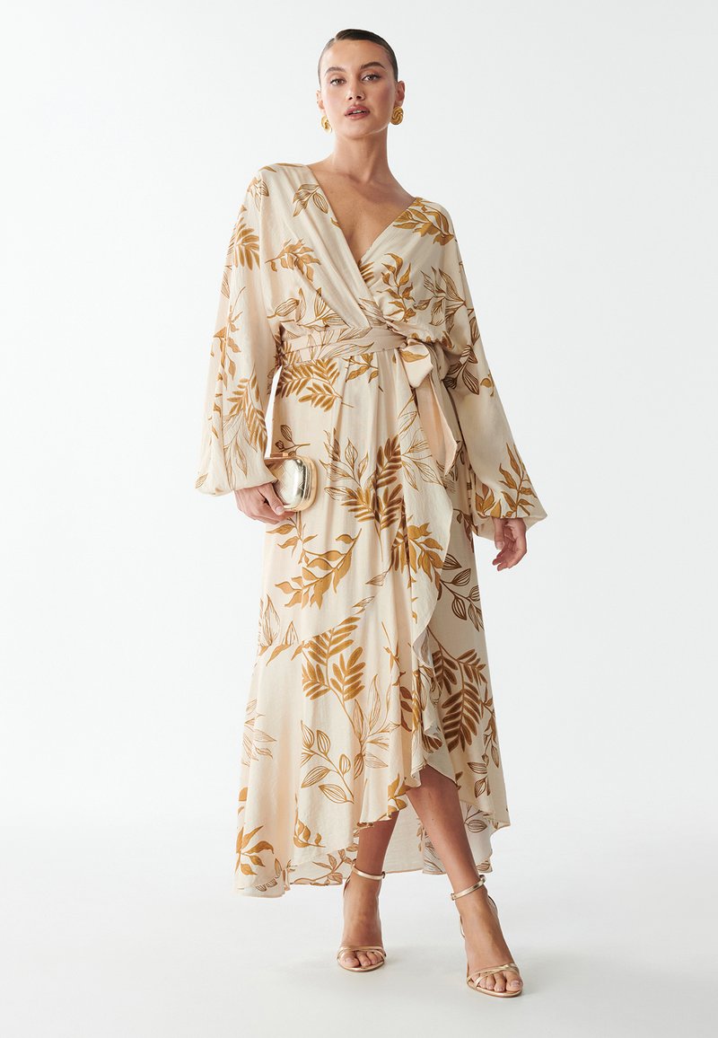 Vrouw in een beige wikkeljurk met bruine bladenpatronen, met een kleine gouden clutch en gouden sandalen en oorbellen.