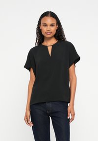 Woman with curly hair wearing a black short-sleeve blouse with a front keyhole detail and dark blue jeans, standing against a white background.