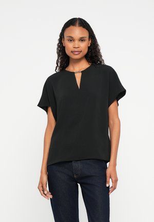 Mulher com cabelo encaracolado vestindo uma blusa preta de manga curta com um detalhe de abertura na frente e jeans azul escuro, apoiada contra um fundo branco.