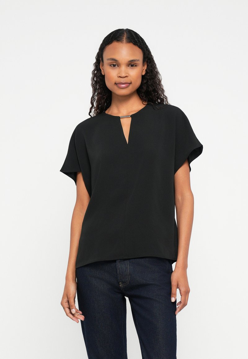 Woman with curly hair wearing a black short-sleeve blouse with a front keyhole detail and dark blue jeans, standing against a white background.
