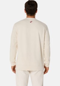 Sweatshirt de couleur crème avec des manches longues, des poignets et un ourlet côtelés. Présente un petit logo rouge sur la zone du cou arrière. Texture douce.