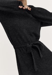 Abito nero in maglia texture con maniche lunghe, cintura annodata in vita e vestibilità rilassata. Il tessuto sembra morbido e leggermente peloso.