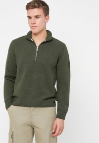 Mladý muž v tmavozelenej polovičnej zip mikine a béžových cargo nohaviciach, stojaci proti jednému bielému pozadiu s rukami v vreckách.