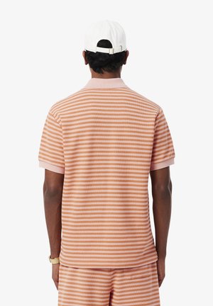 Hombre con camiseta de manga corta de rayas en melocotón y blanco y pantalones cortos a juego, gorra blanca y reloj de pulsera dorado, de espaldas sobre fondo blanco.