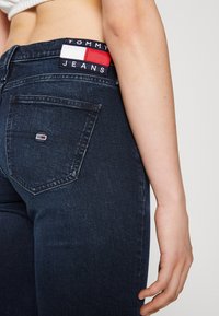 Jeans de mezclilla azul oscuro con corte ajustado; presenta un parche de tela rojo, blanco y azul etiquetado como "TOMMY JEANS" en el bolsillo trasero.