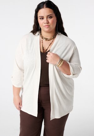 Vrouw met lang donker haar draagt een beige opengebreid vest over een donker shirt en broek, gecombineerd met gelaagde kettingen en meerdere armbanden.