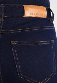 Mörkblå denimjeans med en slät textur, som har kontrasterande orange sömmar och en brun läderetikett präglad med "Noisy May" på baksidan.