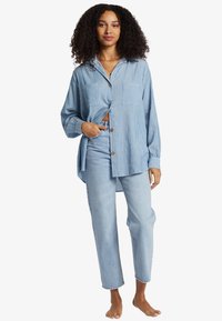 Blue striped button-up shirt with long sleeves, two front pockets; paired with light blue straight-leg jeans. Barefoot.