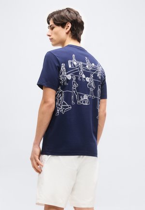 Mlajši moški, oblečen v mornarsko modro majico s belimi risbami ljudi, ki igrajo tenis na hrbtni strani, združeno z belimi hlačami.