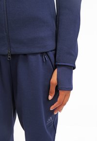 Mörkblå idrottsjacka med dragkedja i texturerat tyg, tumhål i ärmarna och matchande byxor med logotypdetaljer och sidofickor.