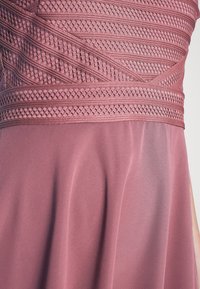 Rosenfarbenes Kleid mit einem strukturierten Mieder und komplexen, gewebten Mustern, das in einen glatten, fließenden Rock übergeht.