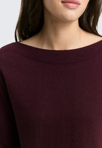 Vrouw met een donkerpaars geribd crewneck sweater, met zichtbare onderkant van haar gezicht en lang donker haar.