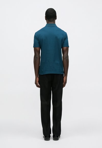 Žalsvo atspalvio polo marškinėliai iš lygaus audinio, su apykakle ir trumpomis rankovėmis, derinami su juodomis tiesiomis kelnėmis. Paprastas dizainas.