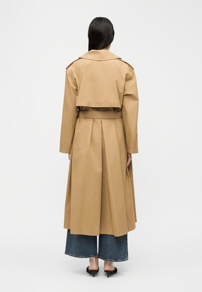 Γυναίκα με μακριά σκούρα μαλλιά, ντυμένη με μπεζ καμπαρντίνα, τζιν παντελόνι με φαρδιά μπατζάκια κομμένο και μαύρα πέδιλα με τακούνι, στέκεται γυρισμένη με την πλάτη.