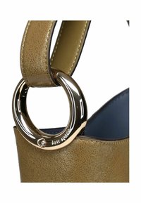 Dettaglio ravvicinato di una tracolla di borsa in pelle color beige attaccata con un anello metallico circolare dorato lucido, che mostra cuciture e texture dettagliate.