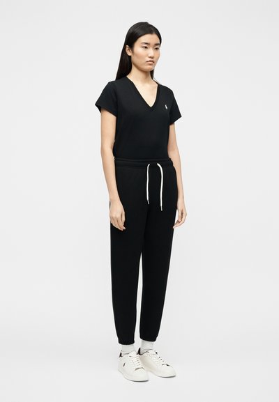 Νεαρή γυναίκα που στέκεται φορώντας μαύρο T-shirt με V-λαιμόκοψη, μαύρα παντελόνια φόρμας με λευκό κορδόνι, λευκές κάλτσες και λευκά αθλητικά παπούτσια.