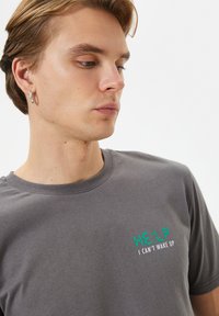Grå bomulds T-shirt med rund halsudskæring og et trykt tekstdesign i grøn og hvid: "HJÆLP JEG KAN IKKE VÅGNE OP" på brystet.