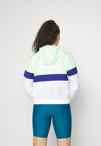 Lekka kurtka z kapturem w paski w kolorze miętowym, białym i granatowym. Posiada elastyczne mankiety i kaptur ze sznurkiem. Noszona z turkusowymi szortami rowerowymi.