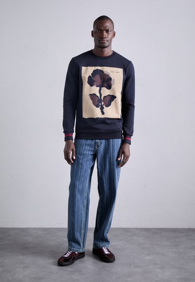 Navy Sweatshirt mit großem beigen Blumenmuster und roten Akzenten, kombiniert mit himmelblauen Jeans mit vertikalen Streifen und burgunderroten Wildlederschuhen.