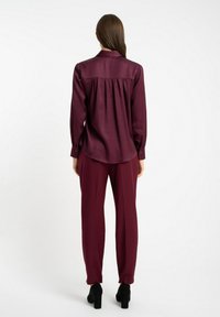 Bordowa jedwabna koszula z ostro zakończonym kołnierzykiem, długimi rękawami i zapinanymi mankietami połączona z dopasowanymi spodniami i czarnymi butami na obcasie.