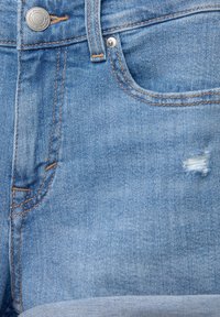 Jean en denim bleu clair en gros plan montrant un bouton, une passants de ceinture, une poche avant et une petite déchirure usée près du bord de la poche.