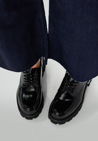 Chaussures noires vernies avec lacets, semelles en caoutchouc épaisses et orteils ronds, associées à un pantalon en denim foncé avec une coupe à la cheville.