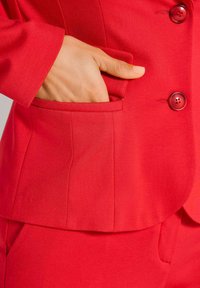 Hand met manicured nagels gestoken in de zak van een rode blazer met bijpassende knopen en zichtbare stofstructuur.