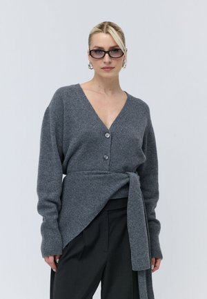 Donna con occhiali da sole scuri, grandi orecchini a cerchio, un maglione grigio con bottoni e pantaloni neri larghi su uno sfondo chiaro e uniforme.