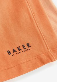 Tela naranja que muestra el texto "BAKER" y "by TED BAKER" en letras negritas de color negro, con costuras visibles y una textura suave.