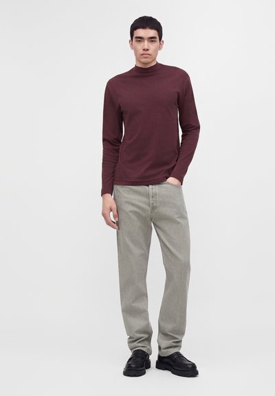 Jeune homme debout portant une chemise rouge foncé à manches longues, un jean gris clair et des chaussures noires sur un fond clair uni.