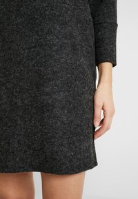 Robe tricotée gris foncé avec une texture douce, des manches longues et un ourlet droit. Main visible près du bas, avec des ongles naturels.