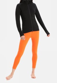 Felpa nera con zip frontale, due tasche laterali e passanti per il pollice, abbinata a leggings arancioni vivaci. Entrambi realizzati con materiali elasticizzati e traspiranti.