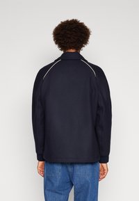 Tmavě modrá bunda s texturovanou látkou, která má sklopený límec a kontrastní bílé švy podél švů na ramenou.