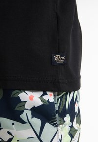 Zwarte katoenen T-shirt met een geweven label waarop "Petrol" in goud staat. Gecombineerd met zwembroek met een bloem- en loofdesign in groen en marineblauw.