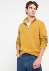 Hosszú ujjú, mustár sárga pulóver fél cipzáros gallérral, bordázott mandzsettákkal és texturált kötött anyaggal. Kék farmernadrággal párosítva.