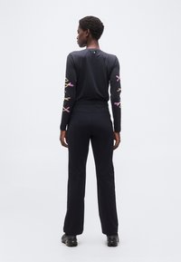 Γυναίκα στέκεται με την πλάτη προς τα εμπρός, φοράει μαύρο μακρυμάνικο πουκάμισο με σχέδια κλαδιών σε ροζ και κίτρινο στα μανίκια και μαύρο παντελόνι, σε λευκό φόντο.