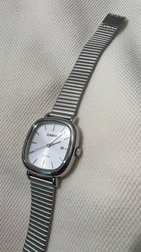 Casio wristwatch with a square silver case, white face, silver hands, date window, and metallic ribbed strap, water-resistant up to 50m.