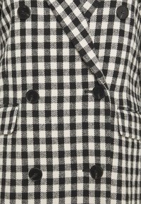 Dubbelbreasted jas gemaakt van zwart-wit geruit stof. Voorzien van gekerfde revers, twee steekzakken aan de voorkant en zwarte knopen.