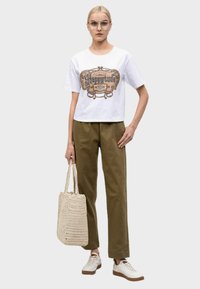T-shirt bianco cropped con stampa grafica, abbinato a pantaloni verde oliva e sneakers bianchi. Una borsa a tracolla intrecciata beige completa l'outfit.
