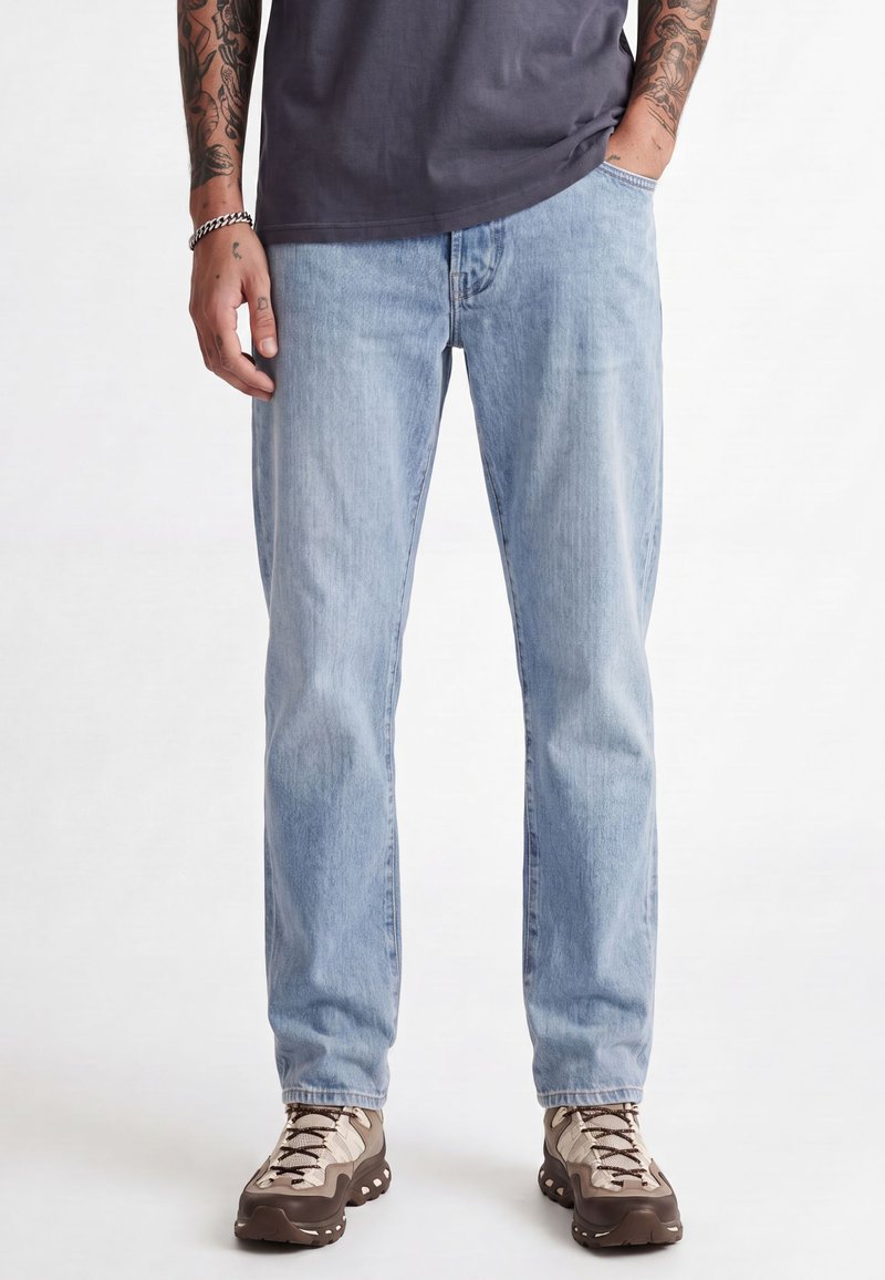 Mand iført lyseblå jeans, mørkegrå t-shirt, synlige armtatoveringer og brune og beige vandresko, der står med den ene hånd i lommen.