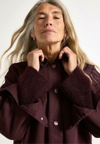 Žena s dlouhými, vlnitými šedými vlasy stojí sebejistě, upravuje si vínově červený kabát s huňatými manžetami, před jednoduchým bílým pozadím.
