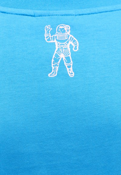 Μπλε βαμβακερό πουκάμισο με λευκό κεντημένο γραφικό αστροναύτη, που στέκεται με ένα χέρι υψωμένο, πάνω σε ομαλή υφή υφάσματος.