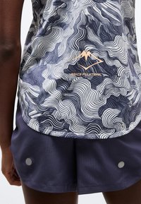 Κοντινό πλάνο ατόμου που φοράει μπλούζα ASICS FUJITRAIL με σχέδιο και σκούρο μπλε σορτς με αντανακλαστικά σημεία στο πίσω μέρος των ποδιών.