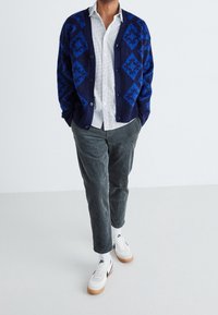 Cardigan blu navale con motivo geometrici, camicia bianca con piccoli punti, pantaloni in corduroy grigi e sneakers bianche con suola in gomma.