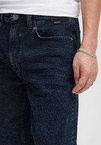 Jeans in denim blu scuro con una finitura testurizzata, dotati di chiusura con bottone e zip, tasche frontali e etichetta con marchio sulla vita.